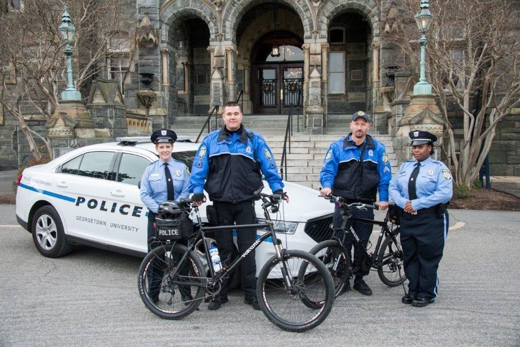 Four GUPD officers stand in front of Healy Hall with a squad car behind them and holding two bicycles.