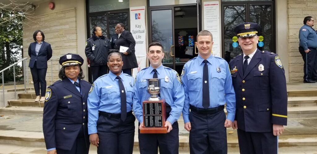 Five GUPD personnel stand for a group photo smiling; the officer in the center holds a trophy.