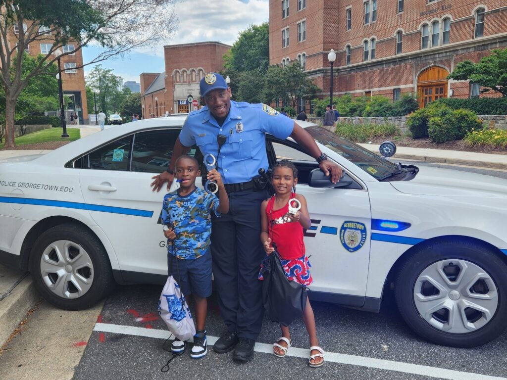 GUPD officer stands in front of squad car with two children posing smiling for a photo.